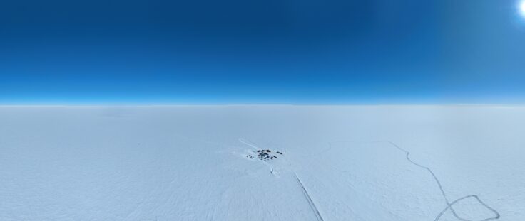 An arial photo of research camp, Little Dome C, on Antarctic ice far in the distance beneath a bright blue sky.