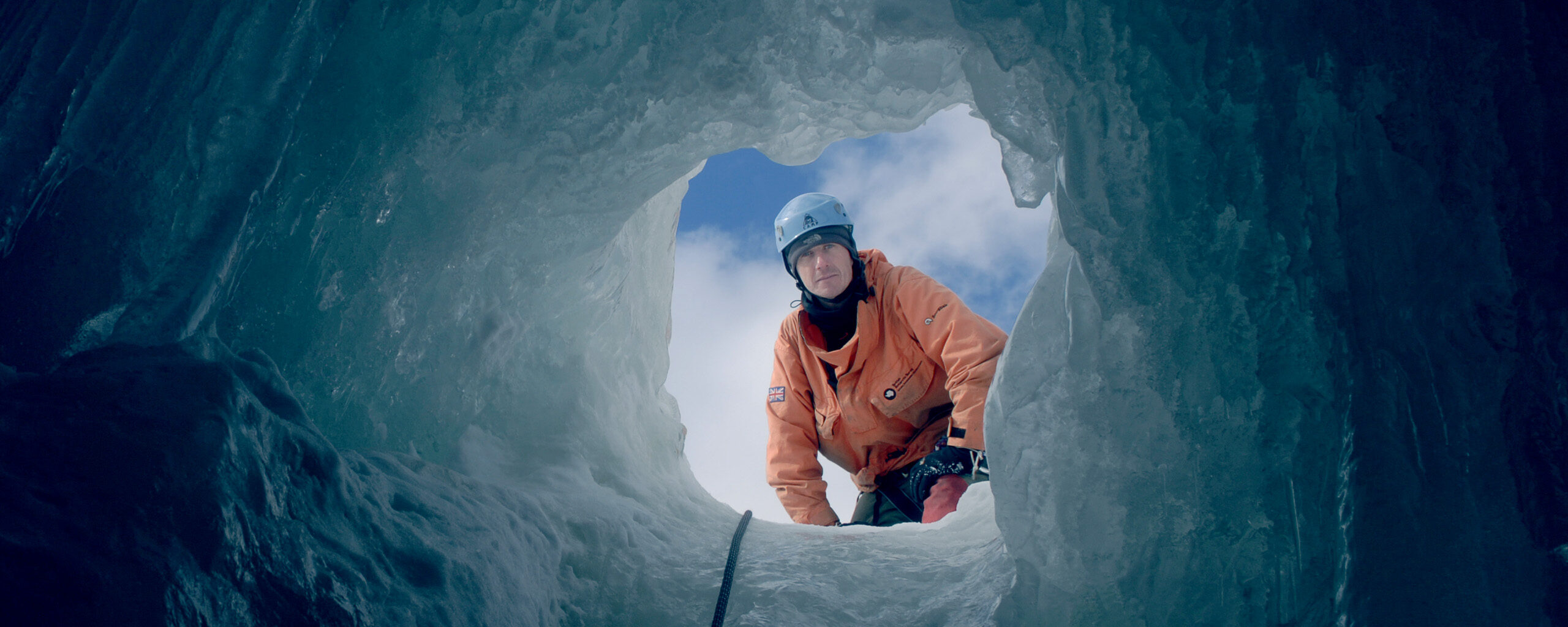 A man wearing safety equipment, attached to a rope looking into the opening of a crevass