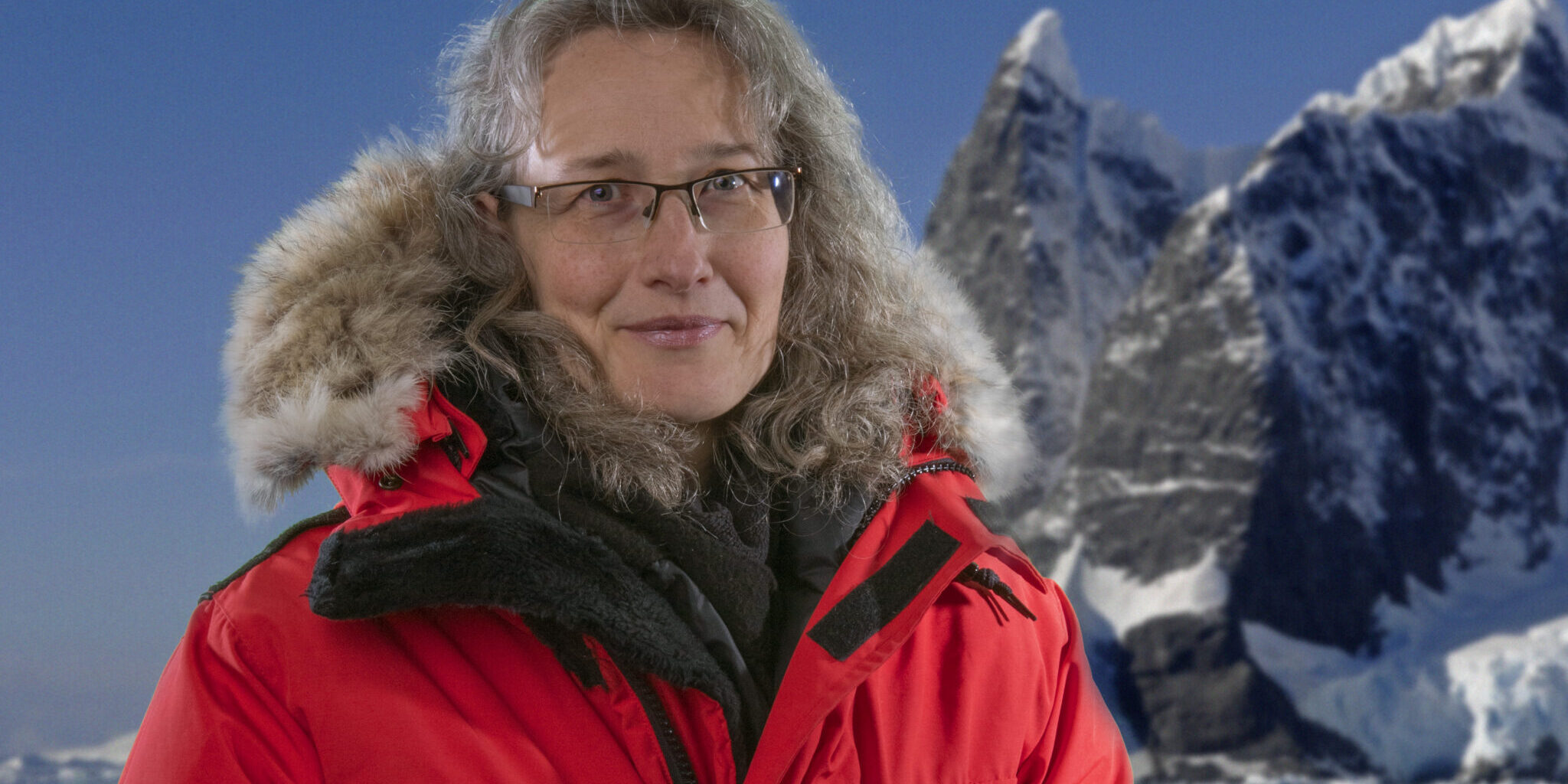 A person that is standing in front of snow covered mountains. They are wearing a bright red parka with a hood.