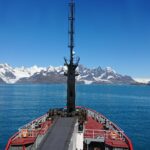 the front of ship in the middle of the sea looking towards snow covered mountains and the sky is blue and cloudless above