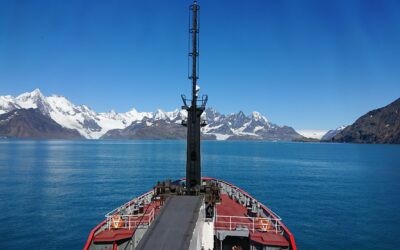 the front of ship in the middle of the sea looking towards snow covered mountains and the sky is blue and cloudless above