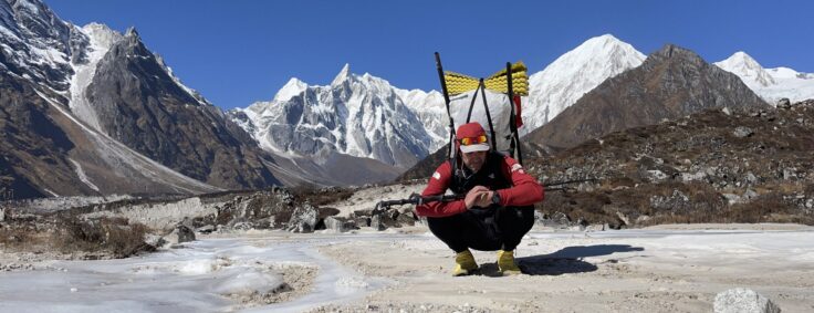 A person crouches to rest in the final moments of a hike to Rara Lake in the Himalaya. They have large a backpack on, in the distance snowy mountains rise above the hilly landscape.