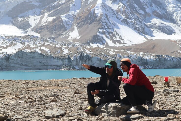 Two people kneel on the rocky outcrop next to a bright blue lake, behind which huge glaciers and mountains rise out of frame. They are looking and pointing into the distance.
