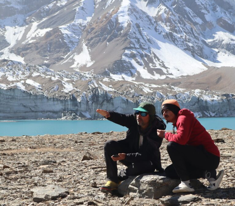 Two people kneel on the rocky outcrop next to a bright blue lake, behind which huge glaciers and mountains rise out of frame. They are looking and pointing into the distance.