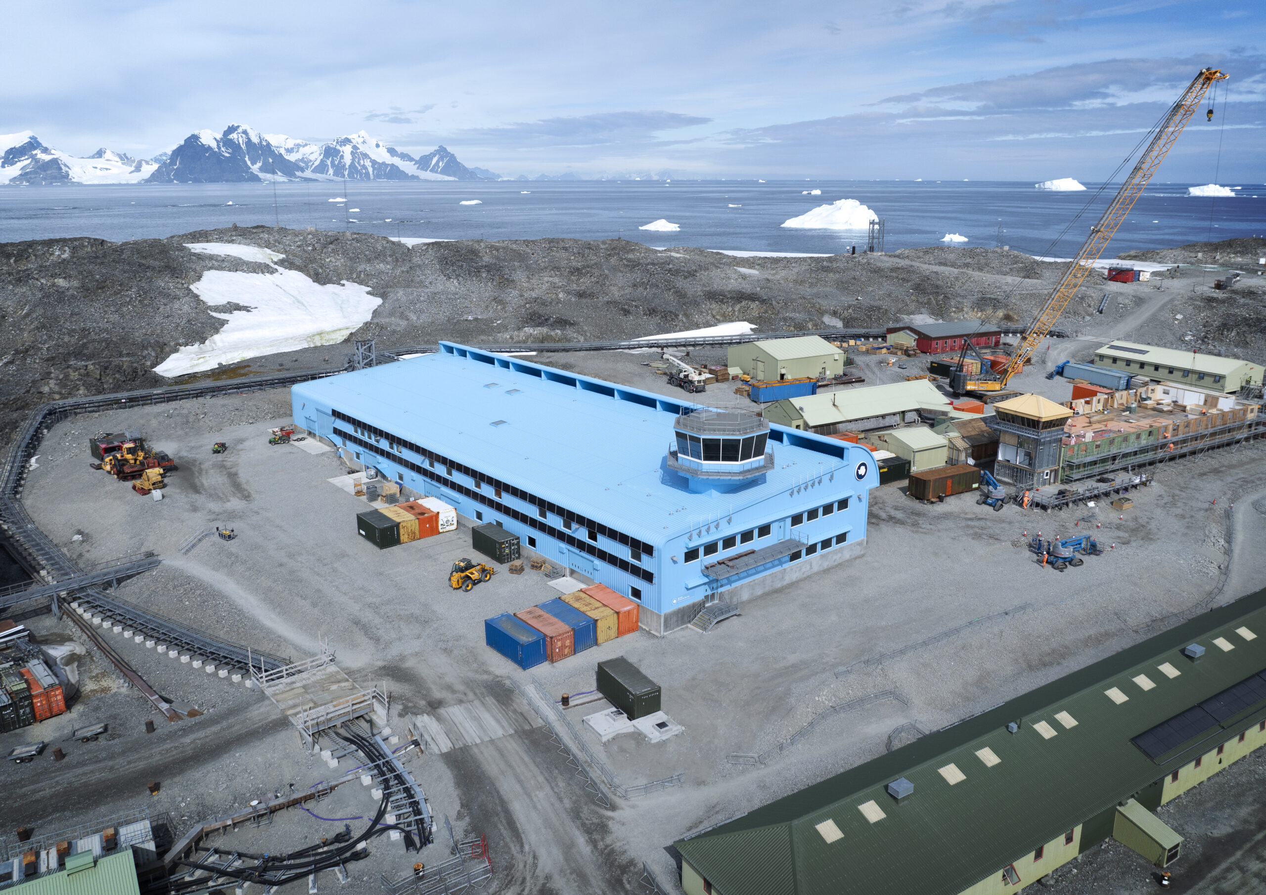 A large blue building with a tower on the top and a curved wind deflector on the side. It's surrounded by construction works and containers and a crane. In the far background there are snow capped mountains and water with icebergs floating.