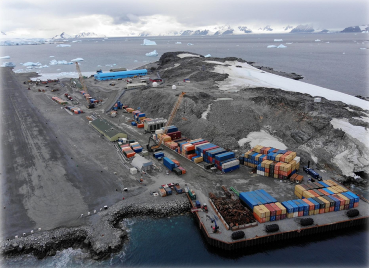 Containers stacked on a wharf. There is a large blue building in the background and snowy mountains. 