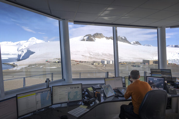 A person at a desk in front of very large windows which give a complete view of the surroundings outside - looking towards a snow covered mountain