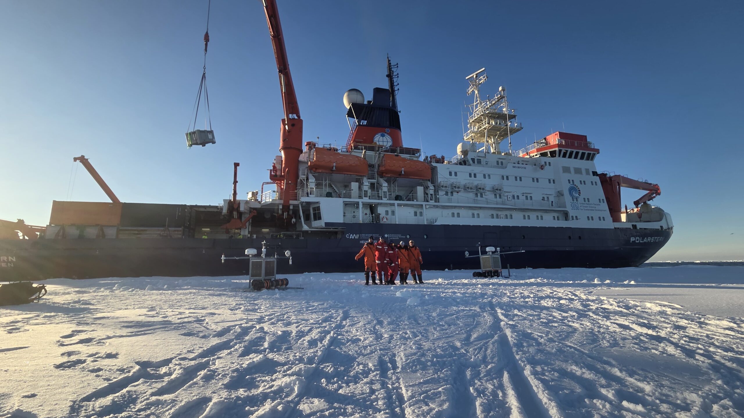 The research ship, the RV Polarstern docked against an ice shelf with scientists posing in the foreground.