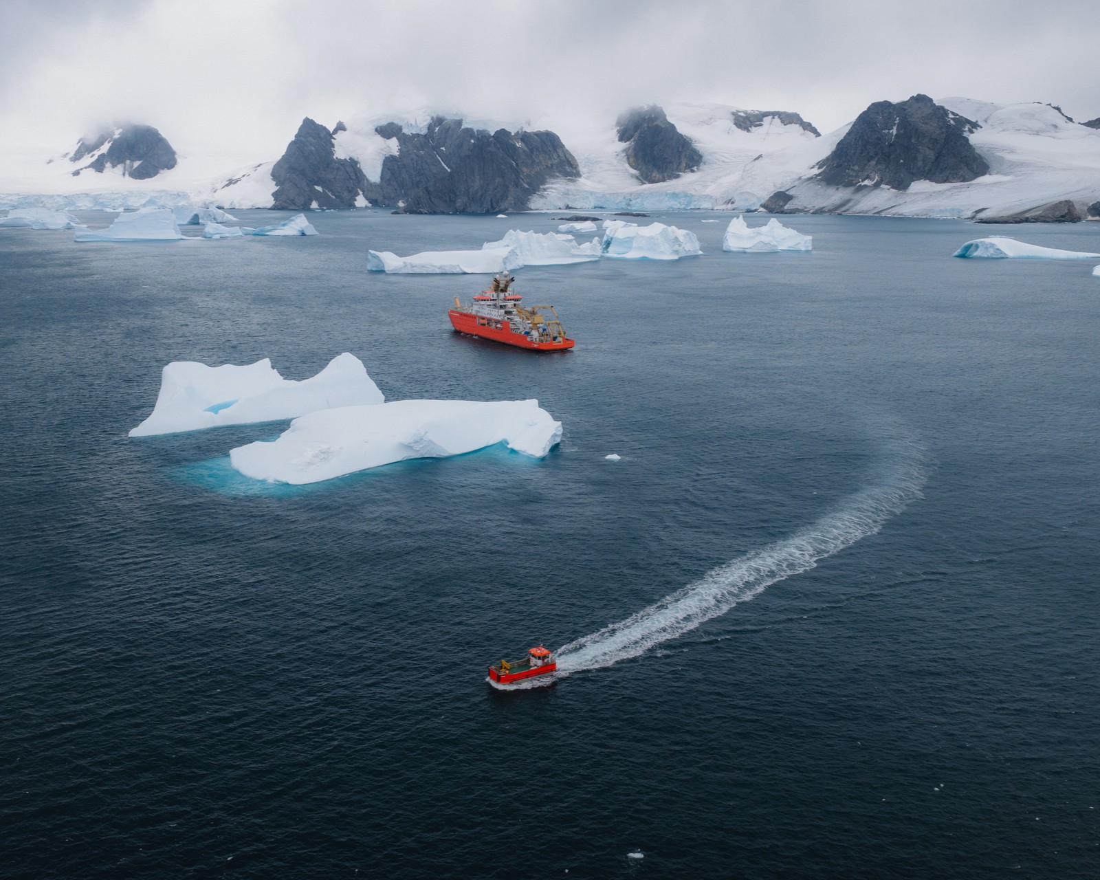 An aerial shot of an iceberg-filled sea in Antarctica. The red research ship Sir David Attenborough sails past an iceberg as the workboat Terror sails in the opposite direction.