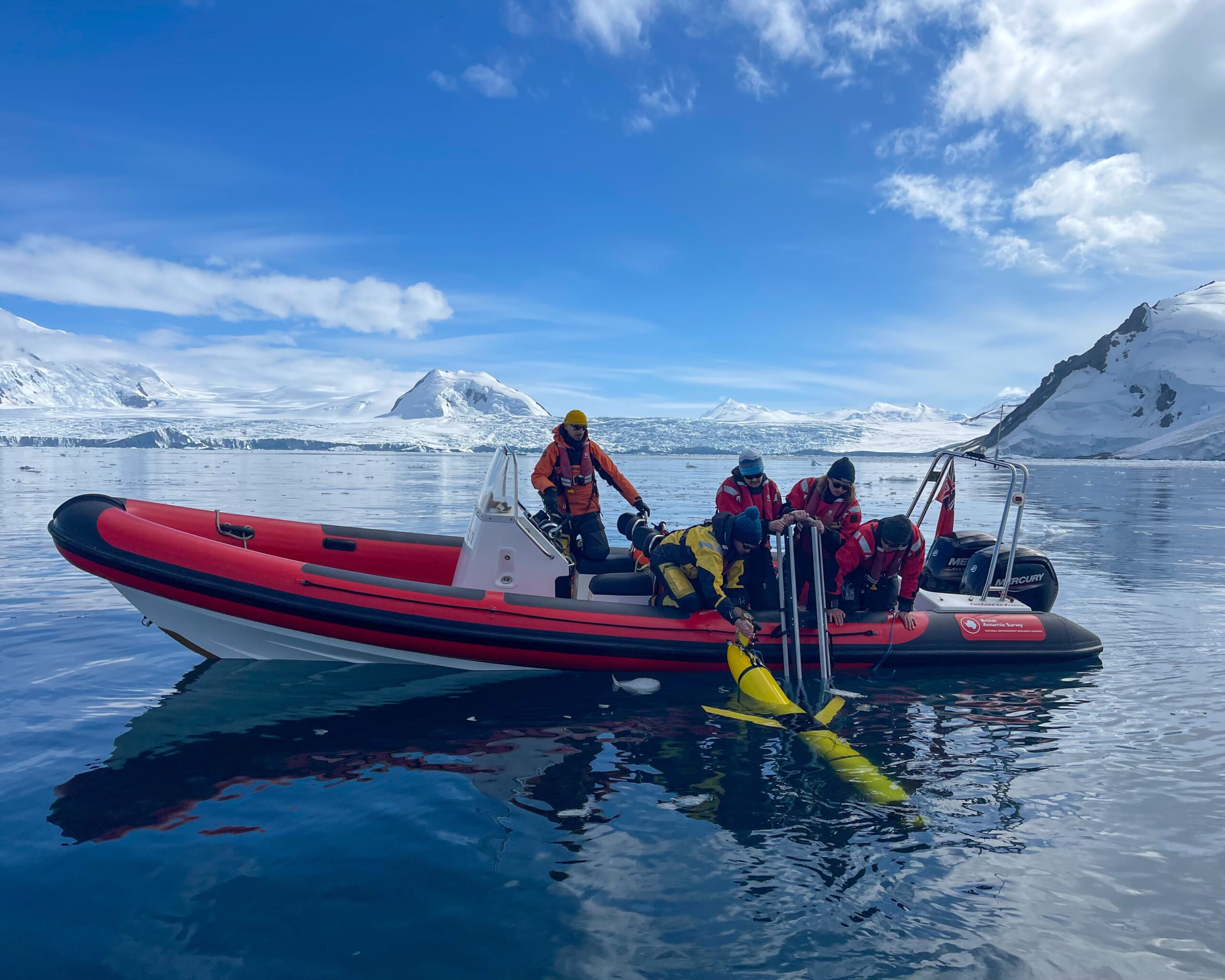 A group of scientists in a boat holding a yellow underwater glider over the side of it at the Sheldon glacier in Antarctica. It's a sunny day and the sea is calm.