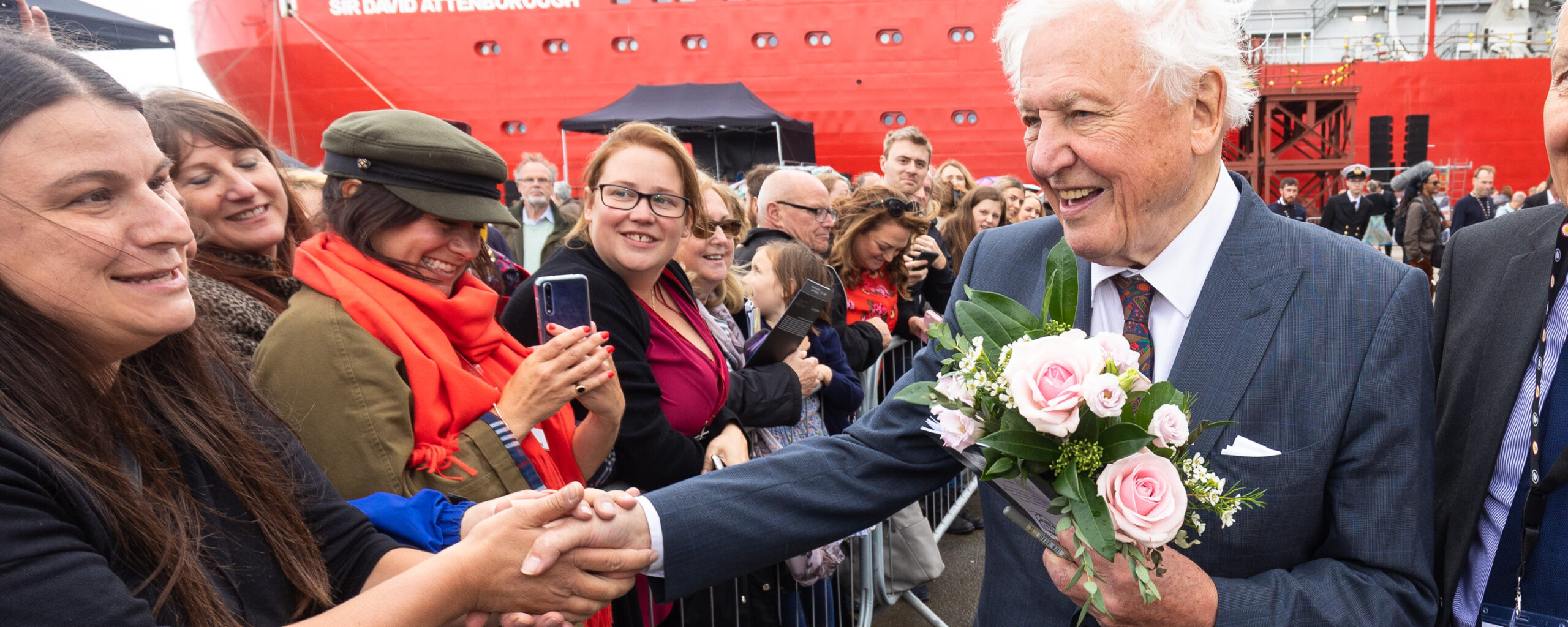 Sir David Attenborough holding flowers as he greets crowds of people.