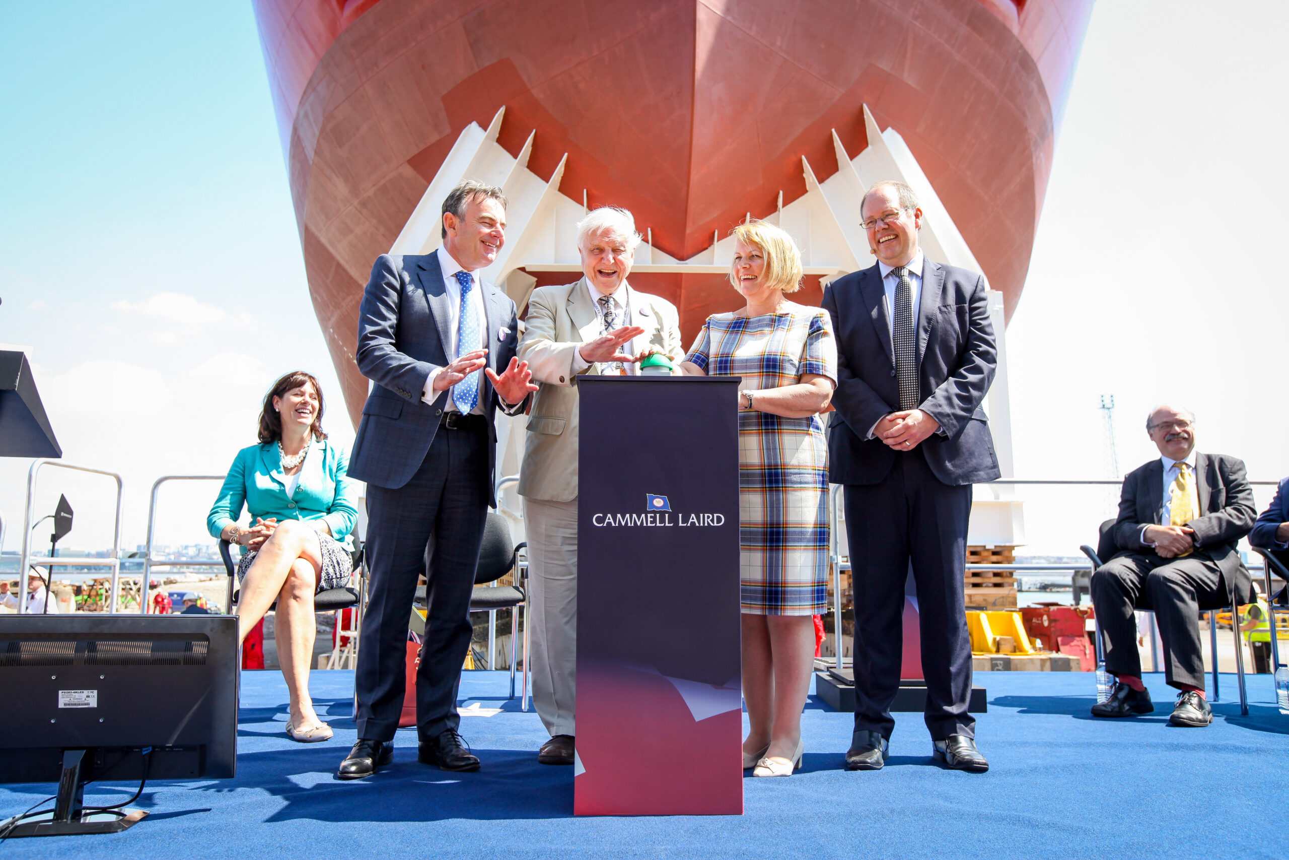 Four people including Sir David Attenborough and Professor Dame Jane Francis people stand around a podium at the bow of the research ship Sir David Attenborough as it sits in a dry dock.