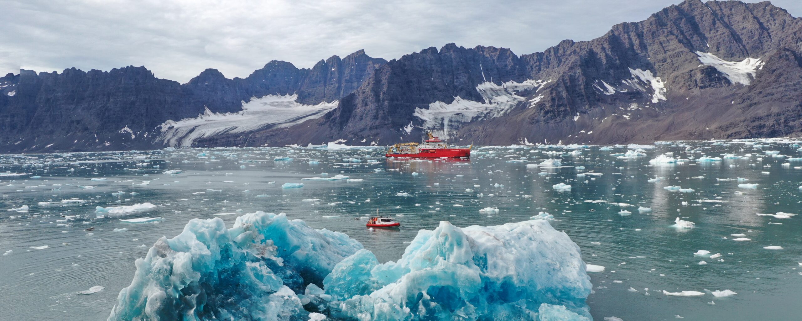A large red ship floats in the distance, with a smaller red boat in front, and an iceberg