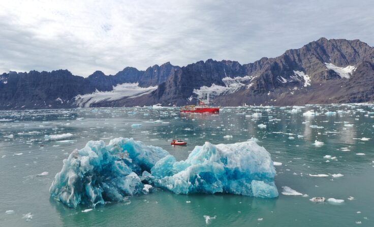 A large red ship floats in the distance, with a smaller red boat in front, and an iceberg