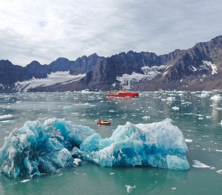 A large red ship floats in the distance, with a smaller red boat in front, and an iceberg