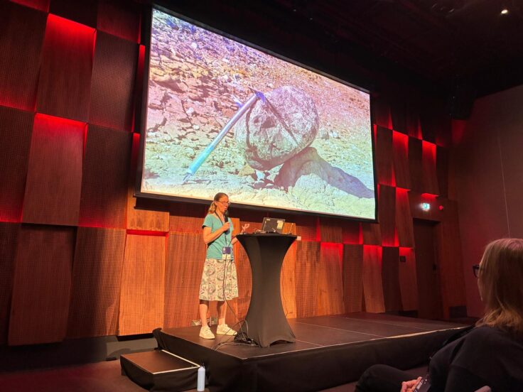 A scientist in front of a large screen showing a rock with a pick axe embedded in it.