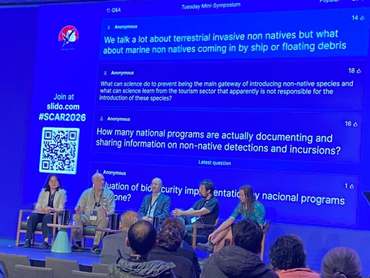 A panel of scientists sit on a stage in front of a blue screen showing audience questions.