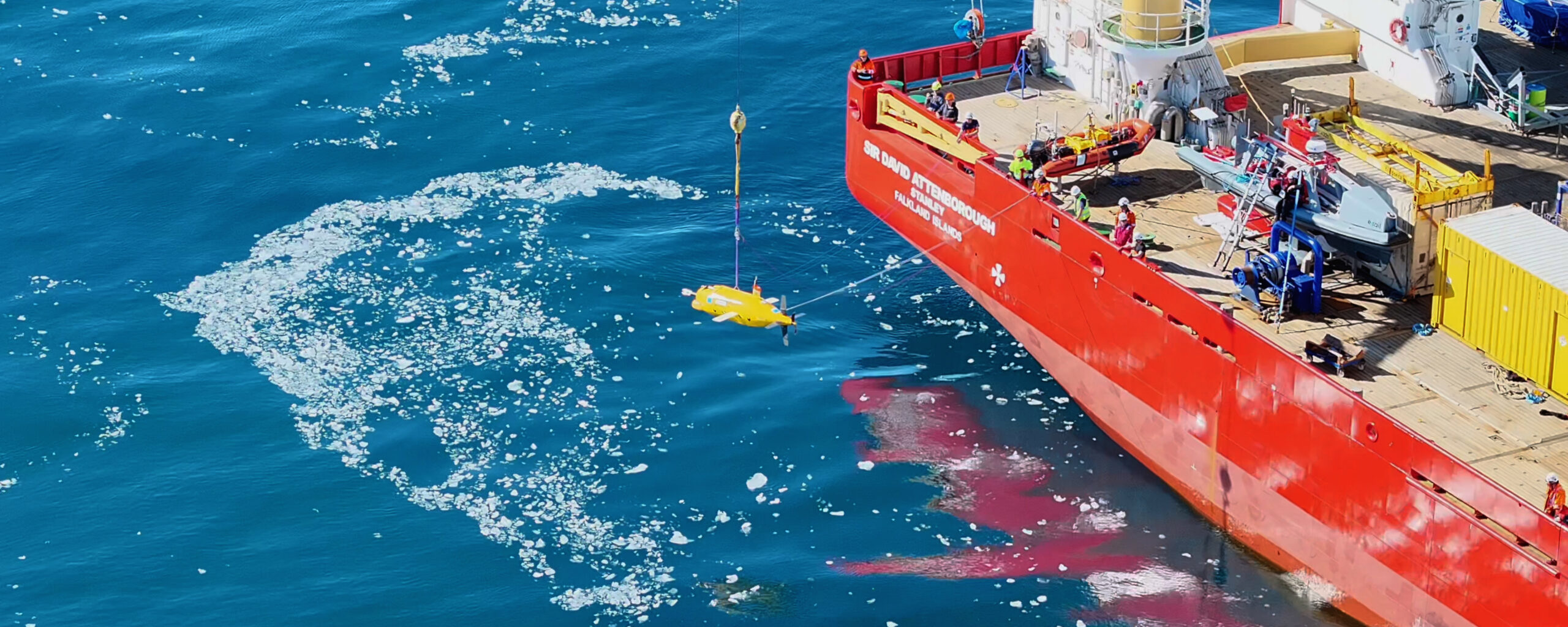 A yellow autonomous submarine being lowered into the ocean from a red ship by a crane.