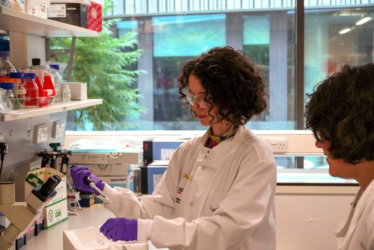 A woman wearing a lab coat and goggles stands in a lab, pipetting a sample