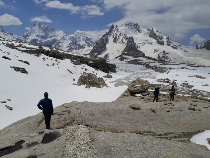 3 people stand by the shores of a frozen mountain lake with snow capped peaks and blue sky.