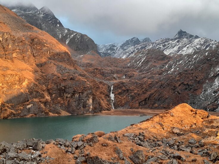 A mountain lake with a waterfall and snow capped peaks in the background.