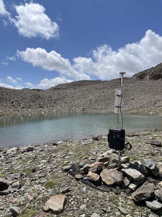 A snowfall sensor on the shores of a stony mountain lake with a blue sky.