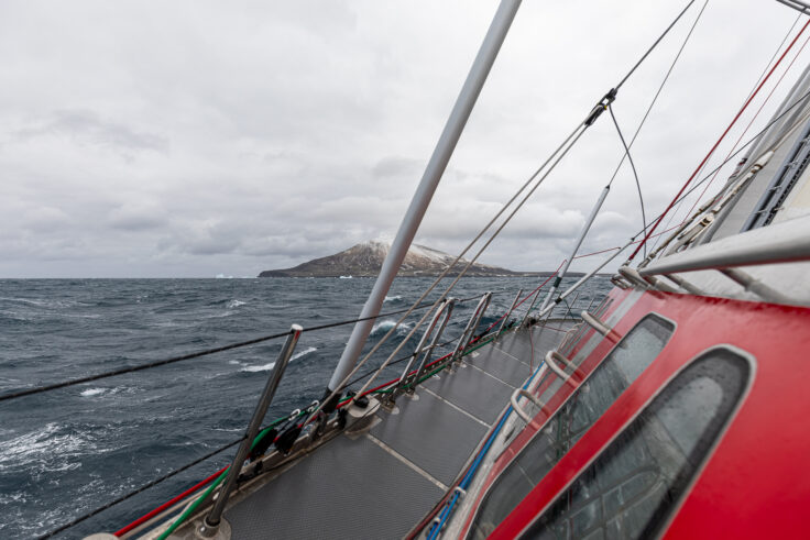 The side of a red yacht on choppy waters, with a volcano in the background.
